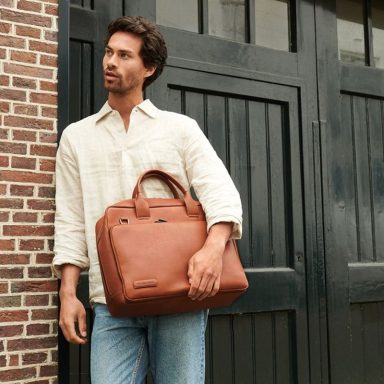 Mann in lässiger Kleidung hält eine braune Tasche, steht vor einer Ziegelmauer.